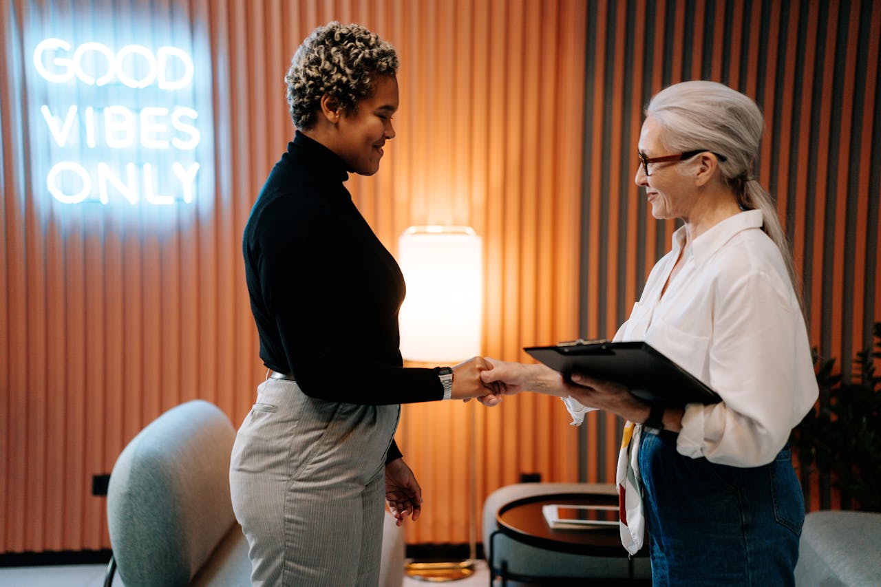 Two diverse businesswomen in a stylish office shaking hands under a Good Vibes Only sign.